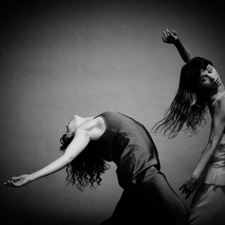 Two dancers in elegant outfits pose in a dynamic movement. The black-and-white shot emphasizes their graceful postures and flowing hair. | © Chris Rogl