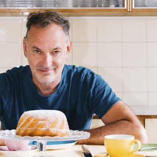 Un homme est assis à une table et regarde un savoureux Gugelhupf. Des tasses et des soucoupes sont dispersées autour de lui sur la table. | © Ingo Pertramer