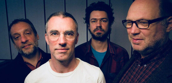 A group of four men stands together in front of a neutral background wall. All have different facial expressions and are wearing different clothes. | © Oliver Potratz
