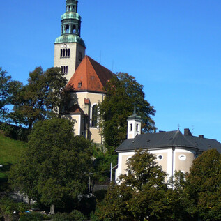 A church with a tall tower and a red roof stands on a hill. Below are some trees and a small building. | © Pfarre Mülln