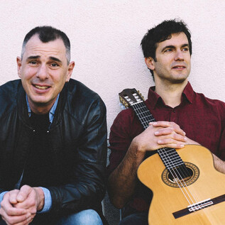 Two men are sitting on a bench, one is smiling, the other has a guitar. They appear relaxed and ready for a musical performance. | © Ingo Pertramer