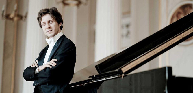A musician in an elegant suit stands next to a grand piano. He has his arms crossed and is looking directly at the camera. | © Marco Borggreve