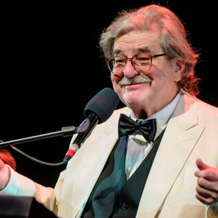 A smiling older man in a white suit and a black bow tie stands at the microphone. He appears friendly and entertaining on stage. | © Karl Satzinger
