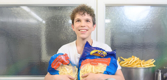 A young man is holding two bags of French fries in his hand. In the background, you can see a kitchen with more fries. | © SLT / Christian Krautzberger