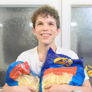 A young man is holding two bags of French fries in his hand. In the background, you can see a kitchen with more fries. | © SLT / Christian Krautzberger
