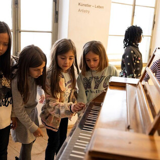 A group of girls is curiously looking at a piano and helping each other. They seem to be having fun exploring the keys. | © Erika Mayer