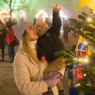Eine Mutter hält ihr Kind in den Armen und zeigt auf einen Weihnachtsbaum. Der Markt im Hintergrund ist stimmungsvoll beleuchtet und hat festliche Dekoration.