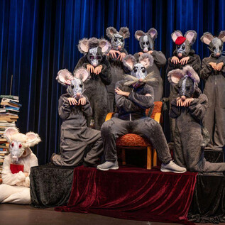 A group of costumed mice stands on a stage, surrounded by a stack of books. In the center, a larger mouse sits on a red throne. | © Leo Fellinger