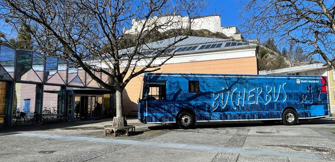 A blue book bus stands in a square next to a modern building. In the background, there is a castle ruin and a clear blue sky. | © Vivien Reichelt
