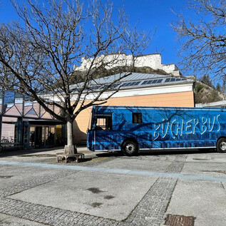 A blue book bus stands in a square next to a modern building. In the background, there is a castle ruin and a clear blue sky. | © Vivien Reichelt