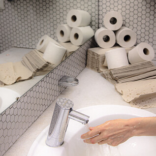 Ein Badezimmer mit einem Waschbecken, einem Wasserhahn und Handtrocknern. Im Hintergrund sind mehrere Rollen Toilettenpapier sichtbar. | © Valentina Schuster