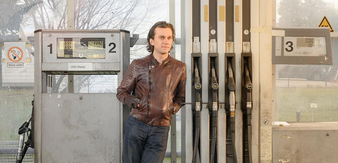 A man is casually standing at a gas station next to the fuel pumps. In the background, faint outlines of trees and a gray sky can be seen. | © SLT / Christian Krautzberger