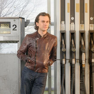 A man is casually standing at a gas station next to the fuel pumps. In the background, faint outlines of trees and a gray sky can be seen. | © SLT / Christian Krautzberger