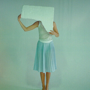 A person is holding a large, rectangular object in front of their face. They are wearing a simple, light-colored skirt and are barefoot on a gentle background. | © Valentina Schuster