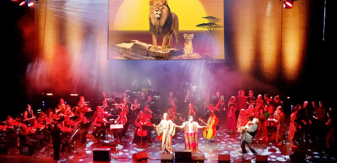 A musical performance on a stage with an orchestra. In the background, there is a large image of a lion and its cubs in front of a sunset. | © COFO Entertainment GmbH & Co.KG