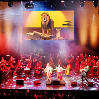 A musical performance on a stage with an orchestra. In the background, there is a large image of a lion and its cubs in front of a sunset. | © COFO Entertainment GmbH & Co.KG