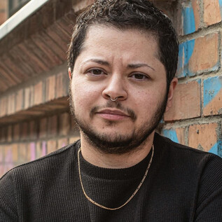 A person stands in front of a brick wall with colorful graffiti. They have a neutral expression and are wearing black outerwear. | © Christine Pichler