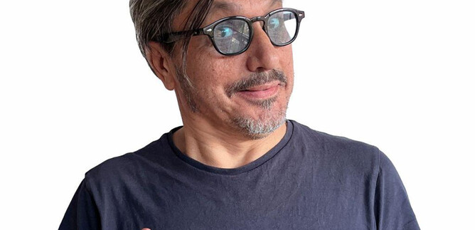 A man with gray hair and glasses smiles and makes a gesture of hope with his hands. He wears a simple black T-shirt and has a friendly expression. | © Kian Noam