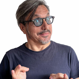 A man with gray hair and glasses smiles and makes a gesture of hope with his hands. He wears a simple black T-shirt and has a friendly expression. | © Kian Noam
