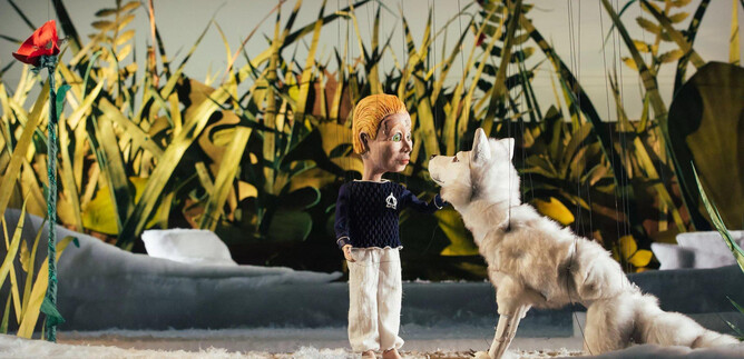 A marionette stands next to a white wolf in a lively, green environment. In the background, tall grasses and red flowers can be seen. | © marco riebler