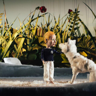 A marionette stands next to a white wolf in a lively, green environment. In the background, tall grasses and red flowers can be seen. | © marco riebler