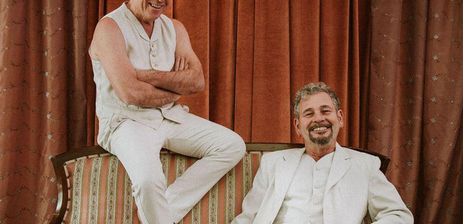 Two men are smiling while posing on a couch. One is wearing a white outfit and the other is dressed in a casual, light ensemble. | © Janna Ramos-Violante