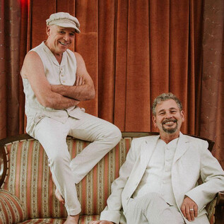 Two men are smiling while posing on a couch. One is wearing a white outfit and the other is dressed in a casual, light ensemble. | © Janna Ramos-Violante