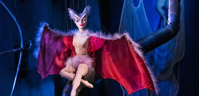 A puppet with bat wings sits on a pole. The background is dark with blue and gray tones. | © georgeye