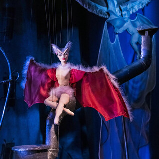 A puppet with bat wings sits on a pole. The background is dark with blue and gray tones. | © georgeye
