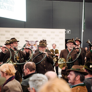 A group of men in traditional costumes is playing hunting horns. They stand in front of an audience and present a musical performance. | © FRBMedia_EmiliaSchloegl