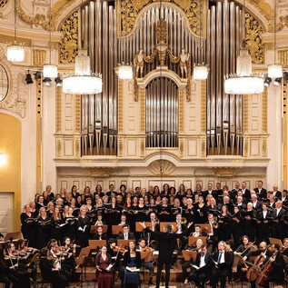 Duże orkiestra i mieszany chór występują na scenie w przepięknej sali. Muzycy i śpiewacy są ubrani w stroje odświętne, a atmosfera jest uroczysta. | © Erika Mayer