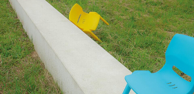 Colorful chairs are standing on a green meadow between gray concrete slabs. The chairs are in various colors like blue, yellow, pink, and green. | © Valentina Schuster