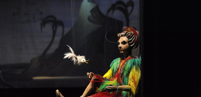 A colorful doll sits on a stage and holds a small bird in her hand. In the background, there are tiered mountains and plants visible. | © Salzburger Marionettentheater