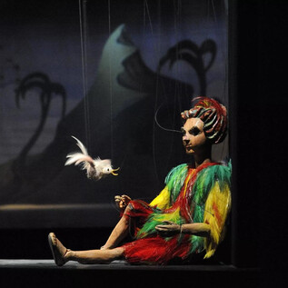A colorful doll sits on a stage and holds a small bird in her hand. In the background, there are tiered mountains and plants visible. | © Salzburger Marionettentheater