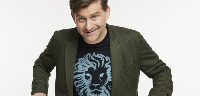 A man with a friendly smile is wearing a T-shirt with a lion motif. He is sitting at a table and has a small, white object in front of him. | © Martina Bogdahn