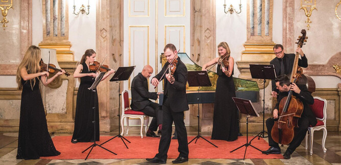 An elegant music ensemble is playing in a festive room. The musicians are dressed in formal attire and are standing in front of music stands. | © Michael Groessinger