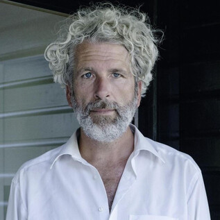A man with curly, gray hair stands in front of a dark wall. He is wearing a white shirt and has a thoughtful expression. | © Gerald von Foris