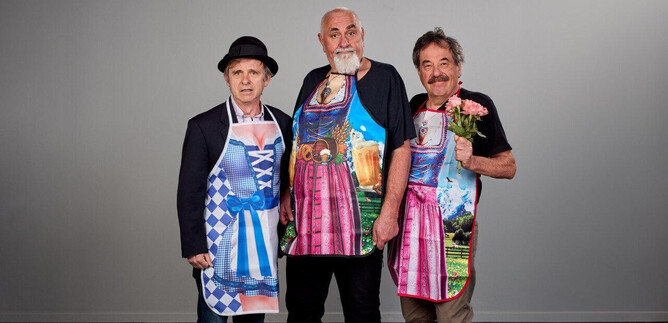 Three men are standing next to each other wearing colorful aprons. They are smiling and posing, while one of them is holding a flower. | © Christian Streili