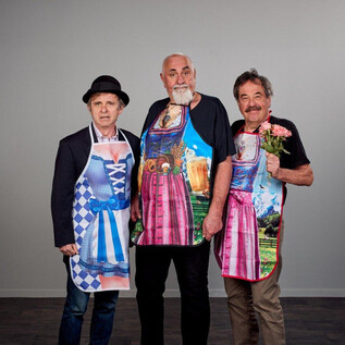 Three men are standing next to each other wearing colorful aprons. They are smiling and posing, while one of them is holding a flower. | © Christian Streili