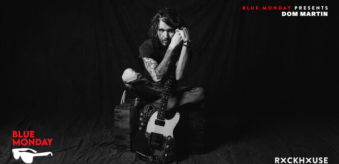 A man is sitting on a crate holding an electric guitar. He is wearing a black outfit and looking thoughtfully at the camera. | © Rob Blackham