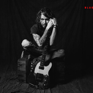 A man is sitting on a crate holding an electric guitar. He is wearing a black outfit and looking thoughtfully at the camera. | © Rob Blackham