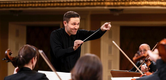 A conductor stands in the foreground and leads the orchestra. The musicians play their instruments in an elegantly designed hall. | © Mozarteumorchester Salzburg