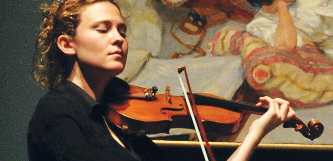 A woman passionately plays the violin in front of a painting. She is wearing a black top and has curly hair. | © Frank Stewart
