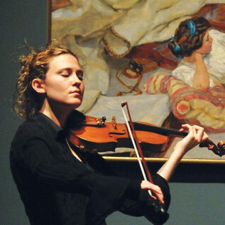 A woman passionately plays the violin in front of a painting. She is wearing a black top and has curly hair. | © Frank Stewart