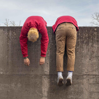Two people are hanging upside down over a concrete wall. They are wearing red and beige clothing and appear relaxed. | © Schauspielhaus Salzburg