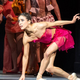 A dancer in a pink outfit moves dynamically on stage. In the background, other dancers in colorful costumes can be seen. | © SLT / Tobias Witzgall