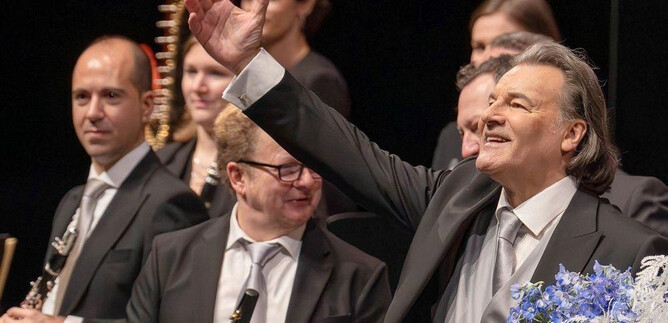 A musician in a black suit smiles and waves to the audience. In the background, other orchestra members with instruments can be seen. | © Christian Schneider