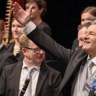 A musician in a black suit smiles and waves to the audience. In the background, other orchestra members with instruments can be seen. | © Christian Schneider