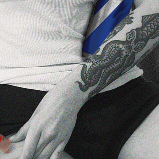 A person injects something into their thigh. The person's arm shows a detailed tattoo and the scene has a monochrome color palette with some colored accents. | © Calixto María Schmutter
