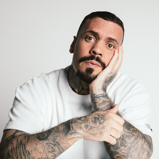 A man with many tattoos looks thoughtfully at the camera. He is wearing a white T-shirt and has a beard. | © Ramiro Simone